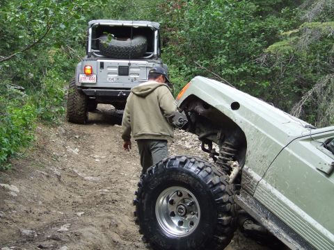 1990 Ford Bronco - Two off-road vehicles navigate a challenging dirt path surrounded by dense forest.