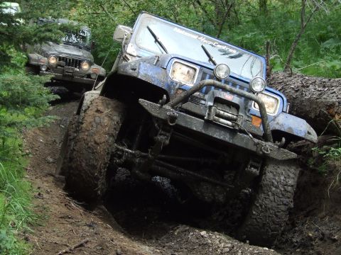 1990 Ford Bronco - A blue Jeep Wrangler tackles a challenging muddy obstacle on an off-road trail, with another Jeep following behind.