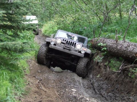 1990 Ford Bronco - Two off-road vehicles tackle a challenging, muddy trail through a wooded area.
