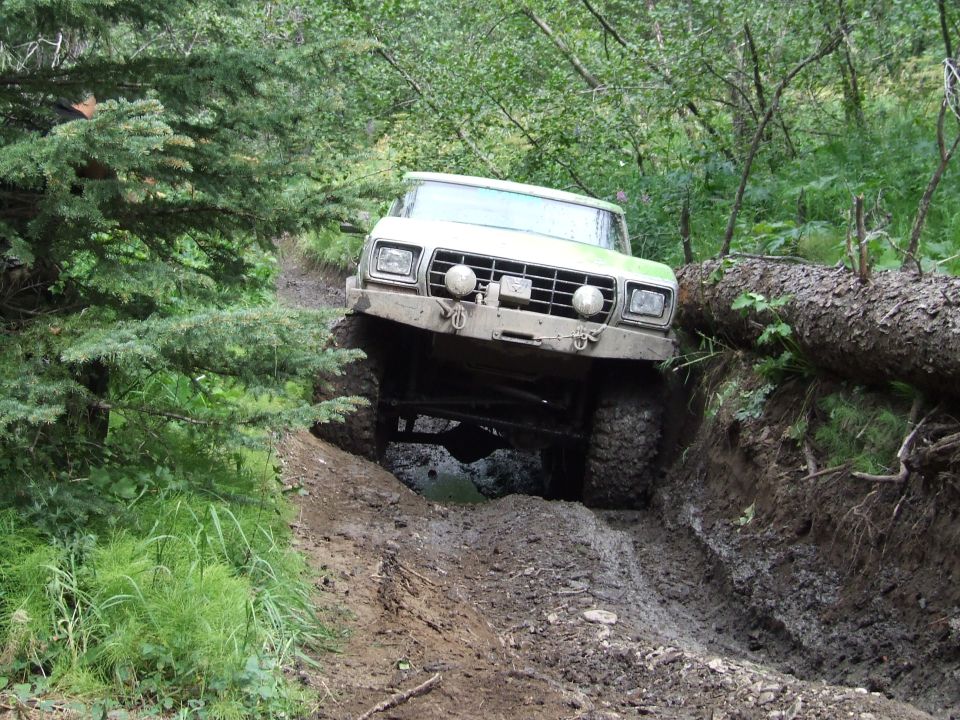Green off-road vehicle with large tires navigating a steep, muddy trail with a fallen tree nearby
