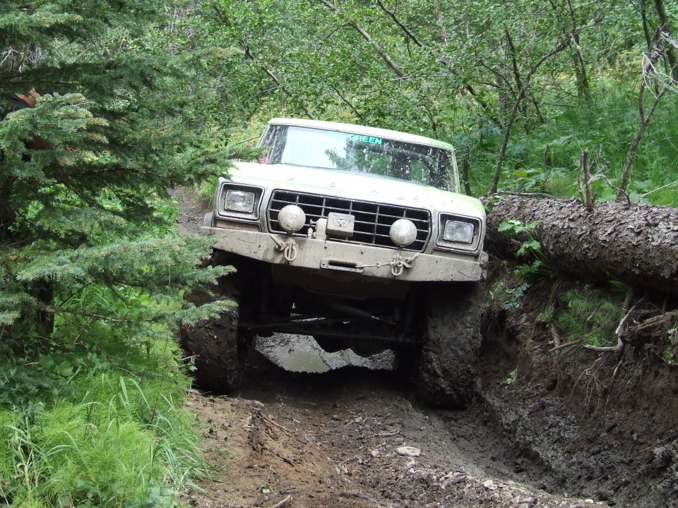 Ford Bronco with large tires navigating a steep, muddy off-road trail with a fallen tree nearby. Text 'GREEN' visible on windshield