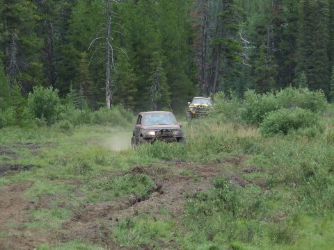 1990 Ford Bronco - Two off-road vehicles navigate a dirt track in a wooded area.