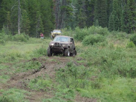 1990 Ford Bronco - Two off-road vehicles navigate a muddy path through a grassy, wooded area.