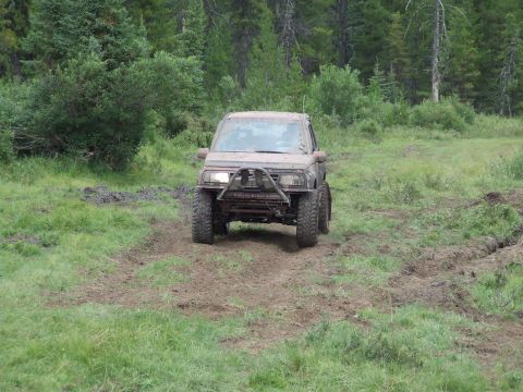 1990 Ford Bronco - Suzuki Samurai navigates a muddy off-road path surrounded by trees.