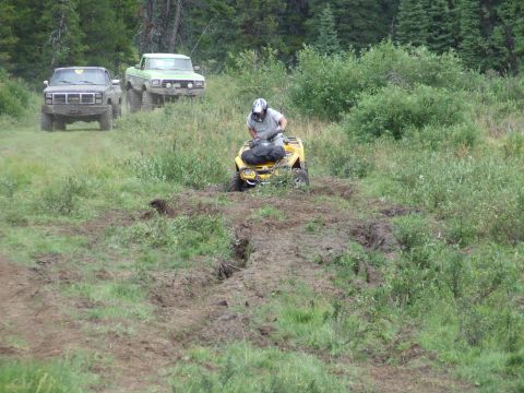 1990 Ford Bronco - Two trucks and an ATV navigate a muddy, grassy terrain during an off-road event.