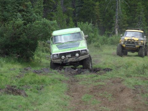 1990 Ford Bronco - Two modified trucks navigate a muddy off-road course surrounded by trees.
