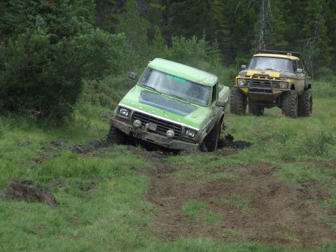1990 Ford Bronco - Two off-road trucks navigate a muddy terrain, with one green Ford pickup truck deeply stuck in the mud.