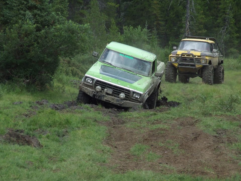 Green Ford pickup truck stuck in mud, front-left wheel deep in mud. Yellow Ford truck behind