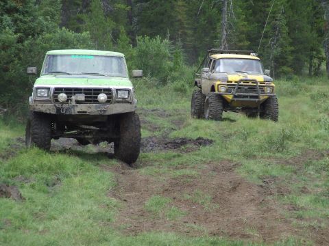 1990 Ford Bronco - Two modified off-road trucks navigate a muddy path through a wooded area.