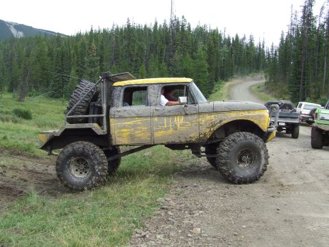 1990 Ford Bronco - A heavily modified pickup truck with oversized tires navigates a dirt road through a forest.