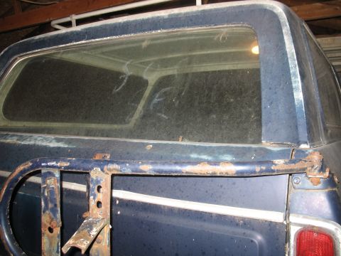 1978 Ford Bronco - The rear of a blue SUV, possibly a Bronco, is shown with a rusty spare tire mount and a white stripe along its side.