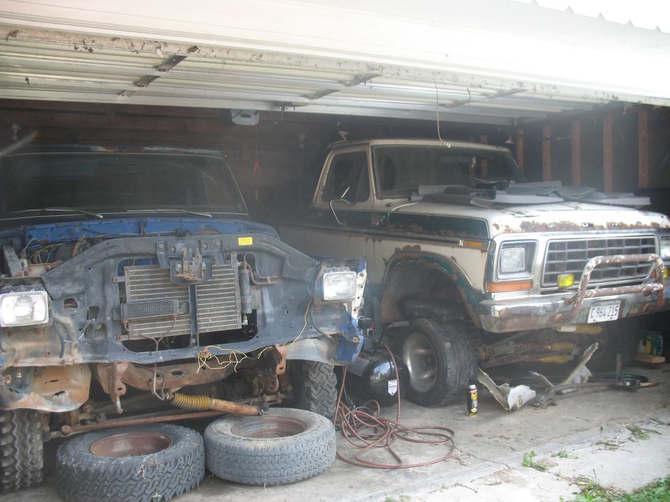 Two Ford trucks, one with front end removed, parked in a garage. The truck on the right appears to be a 1978 Ford Bronco