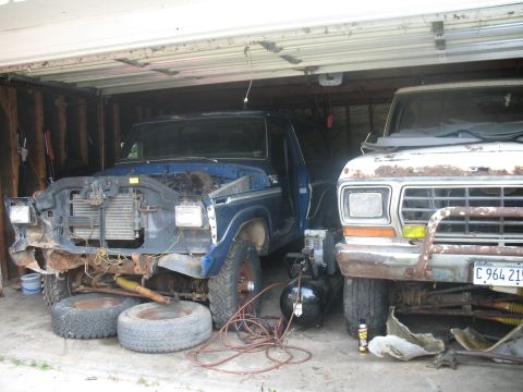 1978 Ford Bronco - Two trucks are parked in a garage, one is blue and missing parts, the other is white and rusty.
