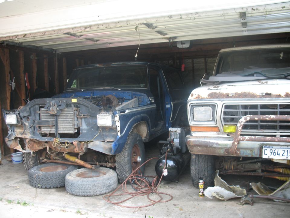 Two old pickup trucks, one blue and partially disassembled, the other white and rusty, parked in a garage. An air compressor sits between them