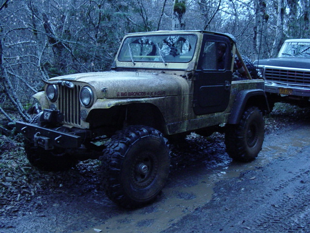 Mud-covered lifted Jeep CJ on large off-road tires in a wooded trail