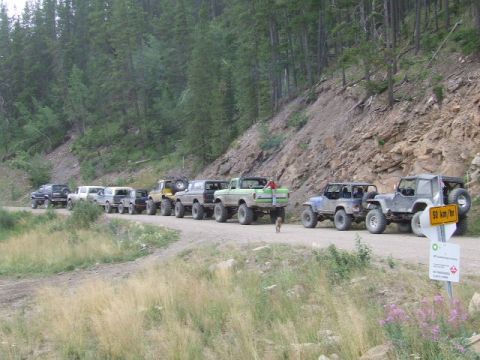 1988 Ford Bronco - A convoy of modified trucks and SUVs travels along a mountain road, with a speed limit sign visible.