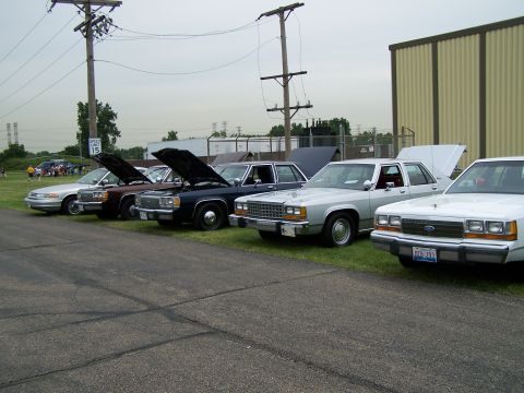 2003 Ford Crown Victoria - Several sedans, including a silver one with its hood open, are parked in a row on a grassy area.