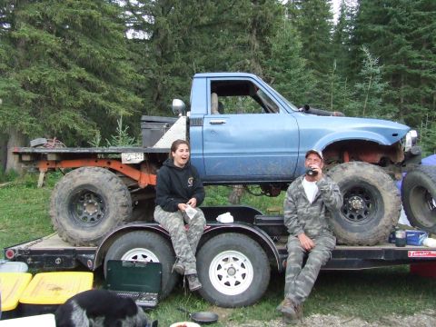 1990 Ford Bronco - Two people relax on a trailer carrying a heavily modified blue pickup truck with large off-road tires.