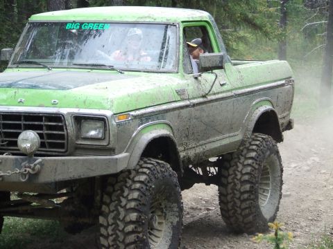 1990 Ford Bronco - A mud-covered green Ford Bronco with "BIG GREEN" on the windshield drives through a wooded area.