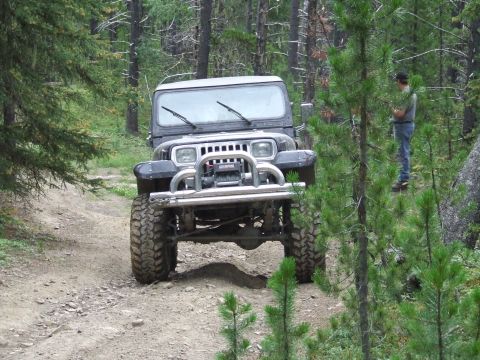 1990 Ford Bronco - A black Jeep Wrangler drives on a dirt path through a forest, with a person standing nearby.