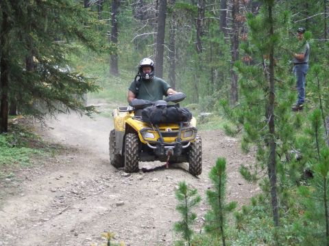 1990 Ford Bronco - A person rides a yellow ATV down a dusty forest road, with another person observing from the trees.