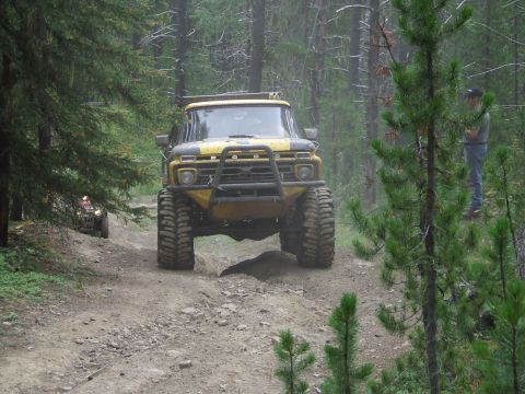 1990 Ford Bronco - A modified yellow truck navigates a wooded trail with another vehicle in the distance.