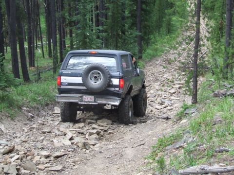 1990 Ford Bronco - A black 1990 Ford Bronco navigates a challenging rocky trail through a dense forest.