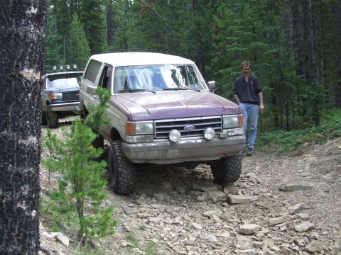 1990 Ford Bronco - A purple and white Ford Bronco navigates a rough, rocky path through a forest with another vehicle behind it.