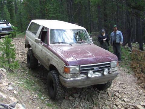 1990 Ford Bronco - A lifted 1990 Ford Bronco navigates a rough, wooded trail with another vehicle visible in the background.