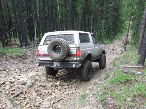 1990 Ford Bronco - A white 1990 Ford Bronco navigates a rough, rocky path through a dense forest.