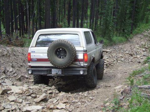 1990 Ford Bronco - A white Ford Bronco navigates a challenging, rocky trail through a forest.