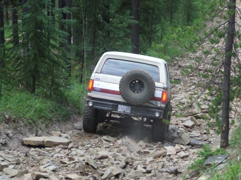 1990 Ford Bronco - A white Ford Bronco navigates a challenging rocky path through a dense forest.