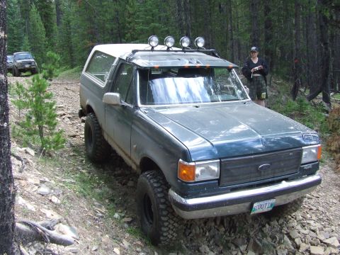 1990 Ford Bronco - A modified Ford Bronco navigates a rocky, wooded trail with another vehicle following behind.