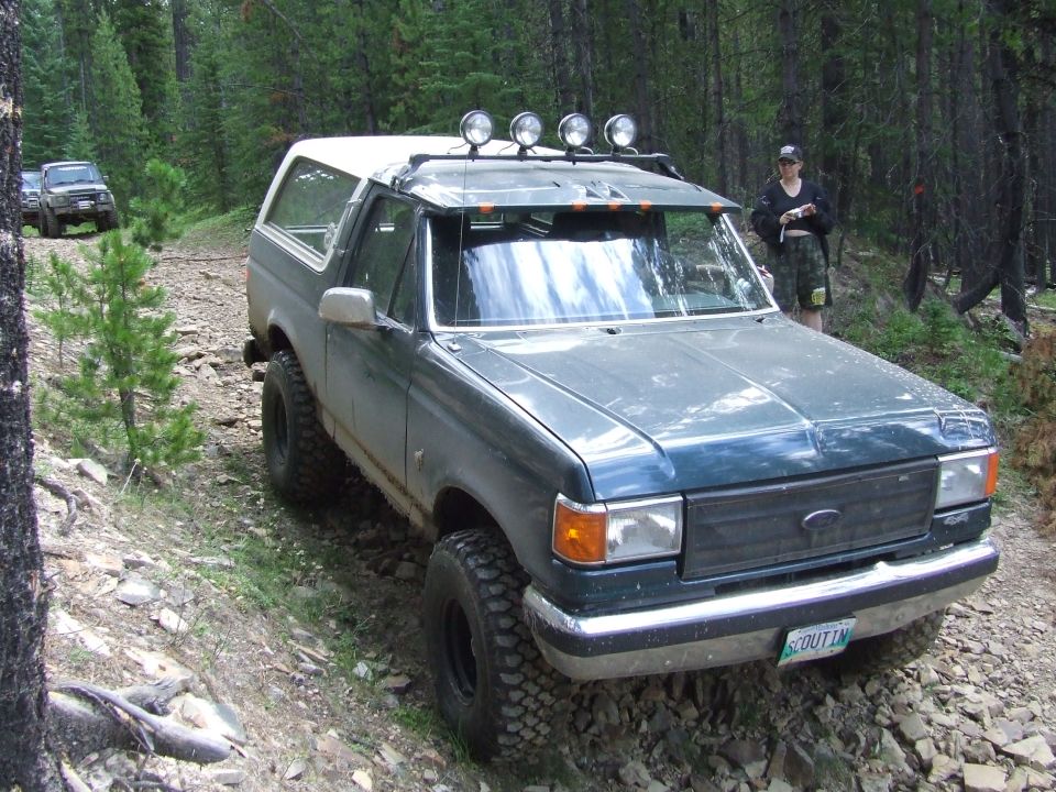 Dark green Ford Bronco with off-road tires on a dirt trail, another SUV behind it. Person standing near the Bronco