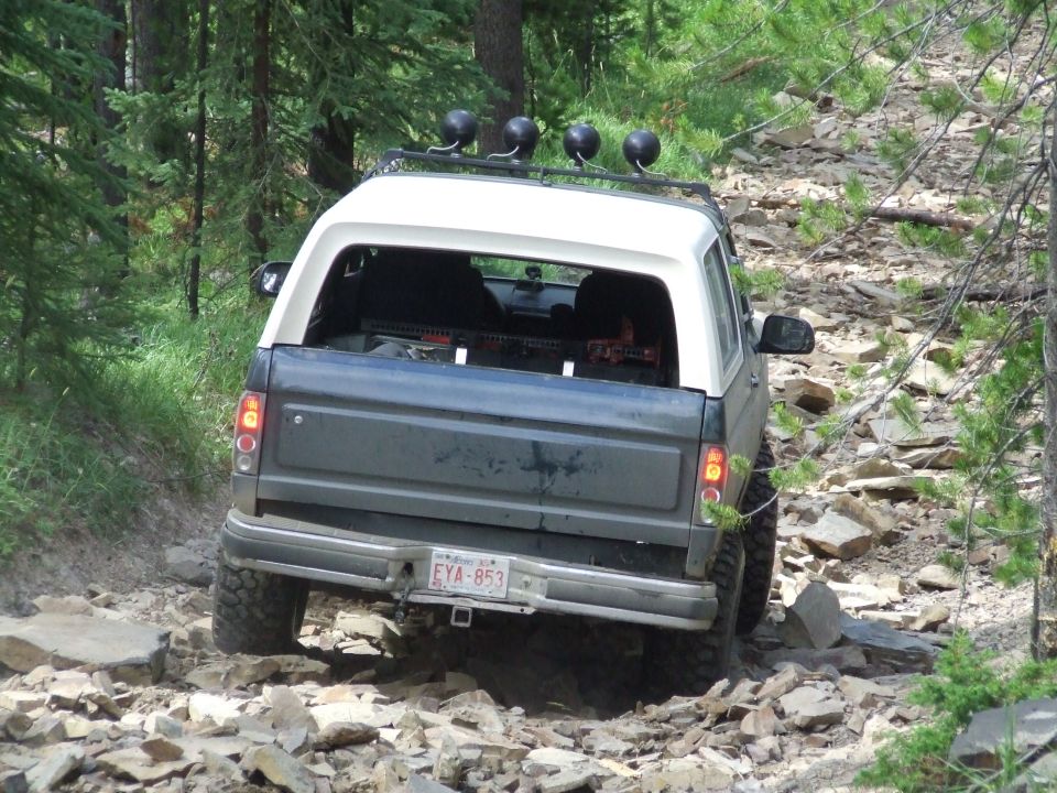1990 Ford Bronco driving on a rocky, wooded trail. License plate EYA-853 is visible