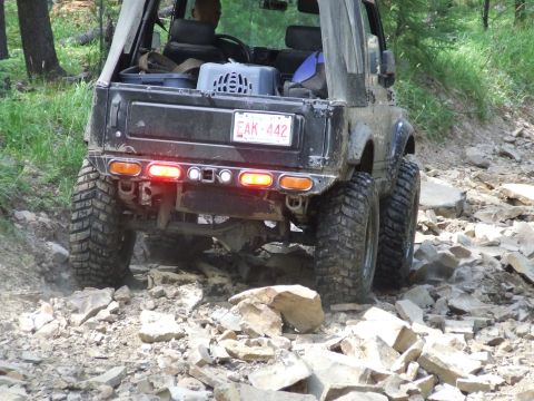 1990 Ford Bronco - Suzuki Samurai navigates a rocky trail, rear view showing off-road tires and lights.