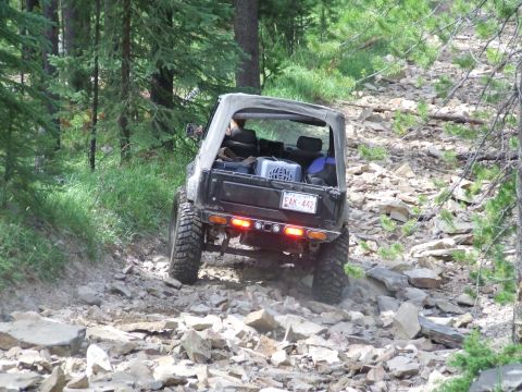 1990 Ford Bronco - A black off-road vehicle navigates a challenging, rocky path through a wooded area.