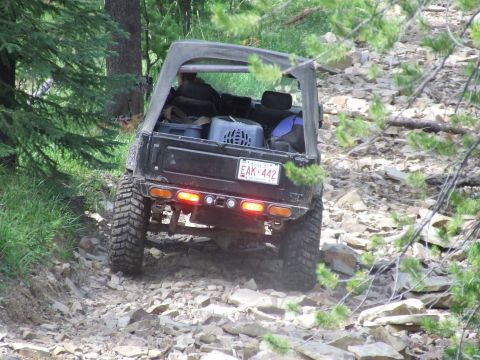 1990 Ford Bronco - A black soft-top off-road vehicle navigates a challenging rocky terrain on a wooded trail.