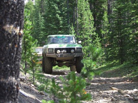 1990 Ford Bronco - A lifted green Ford Bronco navigates a wooded trail, with another vehicle following closely behind.