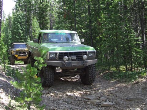 1990 Ford Bronco - Two off-road trucks navigate a wooded, rocky path during an event.