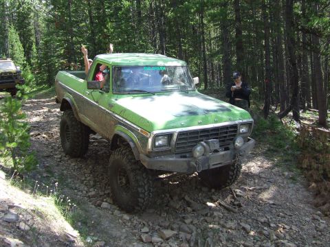 1990 Ford Bronco - A lifted green pickup truck navigates a rocky trail in a forest, with passengers waving from the cab.