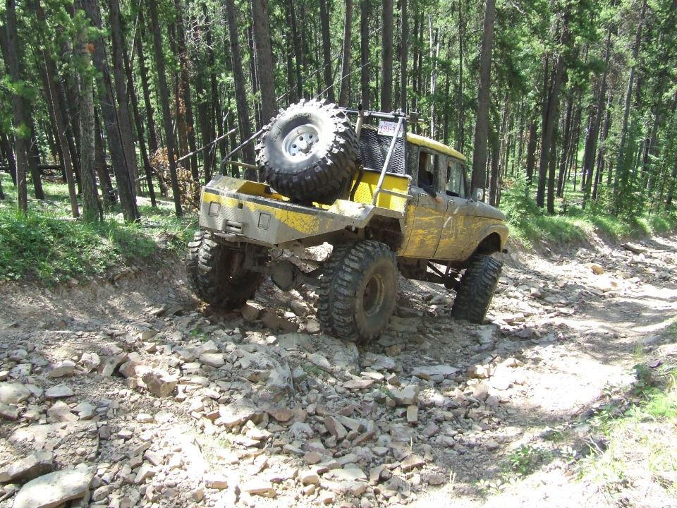 Yellow custom off-road truck with large tires and spare tire on back, driving on a rocky forest trail