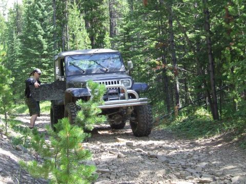 1990 Ford Bronco - A person stands by the open door of a Jeep Wrangler on a wooded, rocky path.