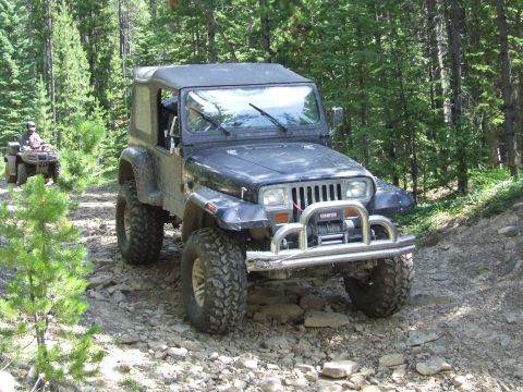 1990 Ford Bronco - A dark blue Jeep Wrangler navigates a rocky forest trail, with an ATV visible in the distance.