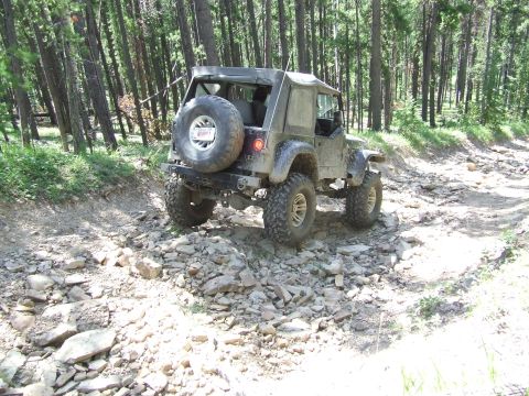 1990 Ford Bronco - A lifted Jeep Wrangler navigates a rough, rocky path through a dense forest.