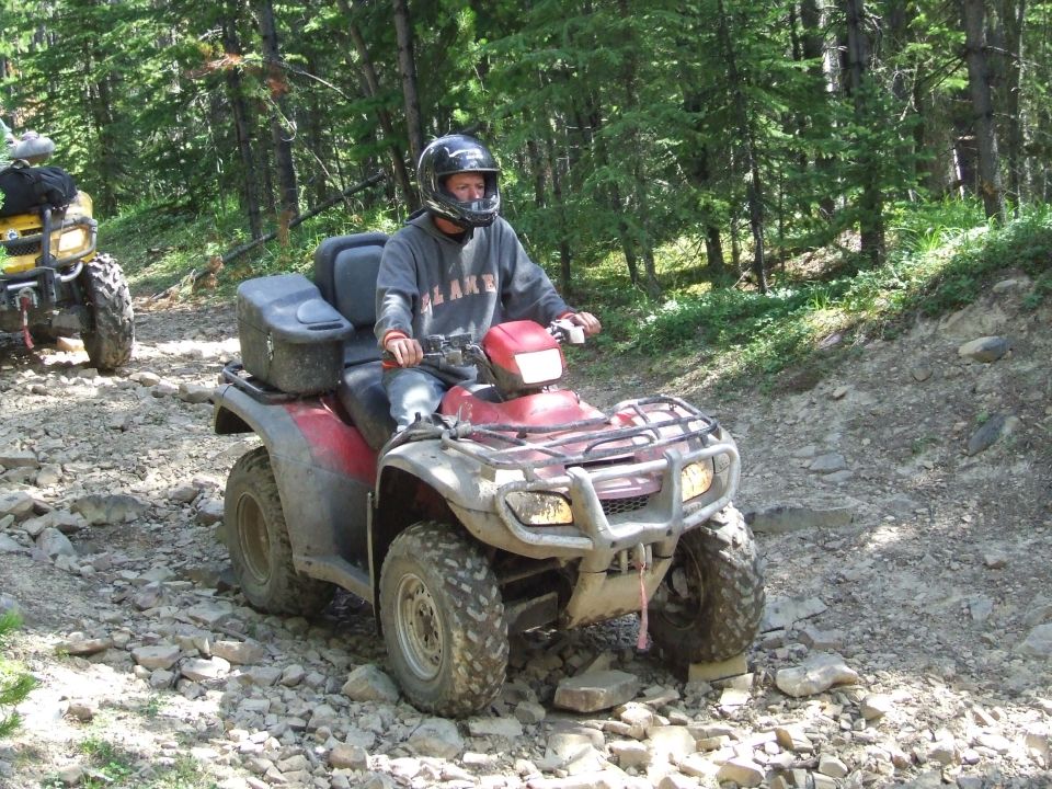 Person riding a red ATV on a rocky path in a forest. Another yellow ATV is visible behind