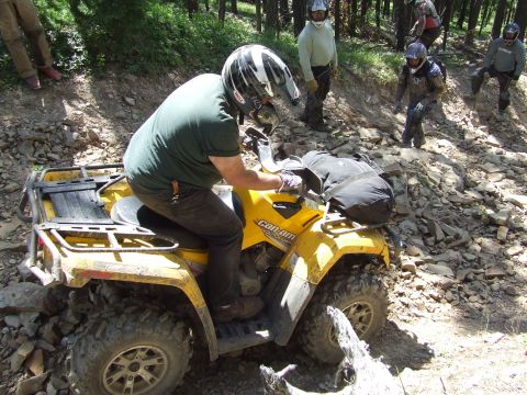 1990 Ford Bronco - A rider navigates a yellow all-terrain vehicle over a rocky, wooded trail with others observing.