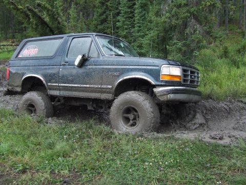 1990 Ford Bronco - A dark blue Ford Bronco navigates a muddy trail, its tires deep in the muck.