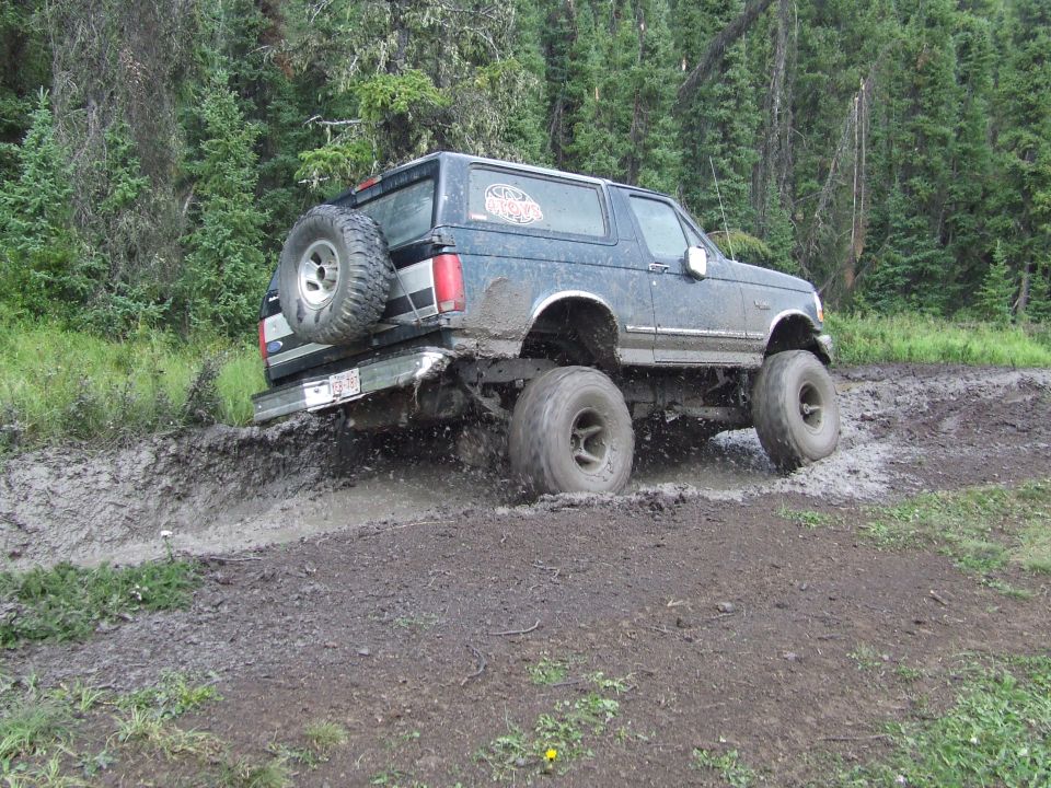 Blue 1990 Ford Bronco driving through mud with large tires and spare on back