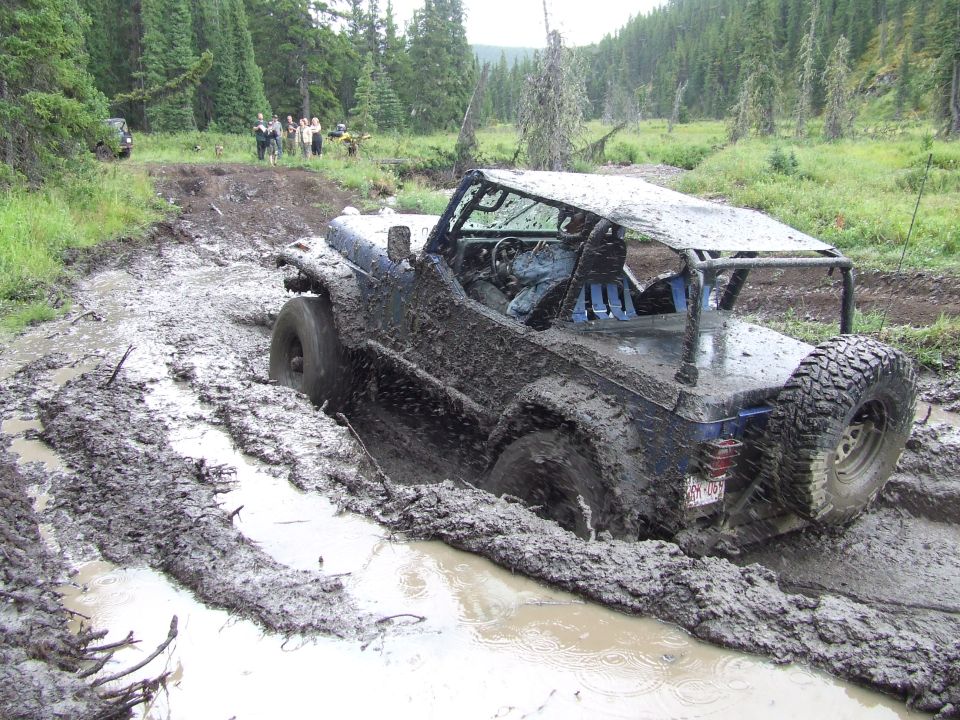 Muddy off-road vehicle stuck in a deep mud pit on a trail. Another vehicle is visible in the background